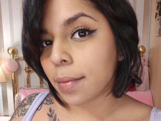 Chili with tan skin and black shoulder-length hair, wearing a white top, has black eyeliner, small pink lips, and a tattoo on her shoulder, looking directly at the camera, slightly smiling.