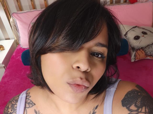 Chili con el pelo negro corto, ojos marrones y tatuajes, acostado en una cama, boca arriba, recibiendo sexo oral de una persona invisible.