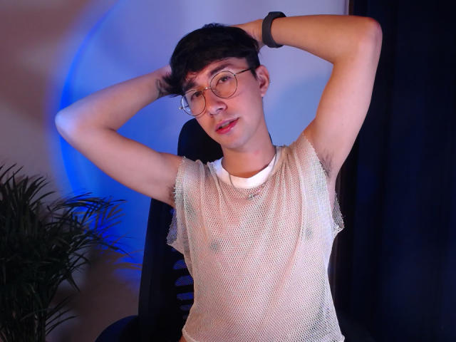 OliverHot69, Asian guy with glasses, short black hair, wearing a mesh white shirt, arms raised, black wristband, plant on left, blue light on wall, black curtain on right.