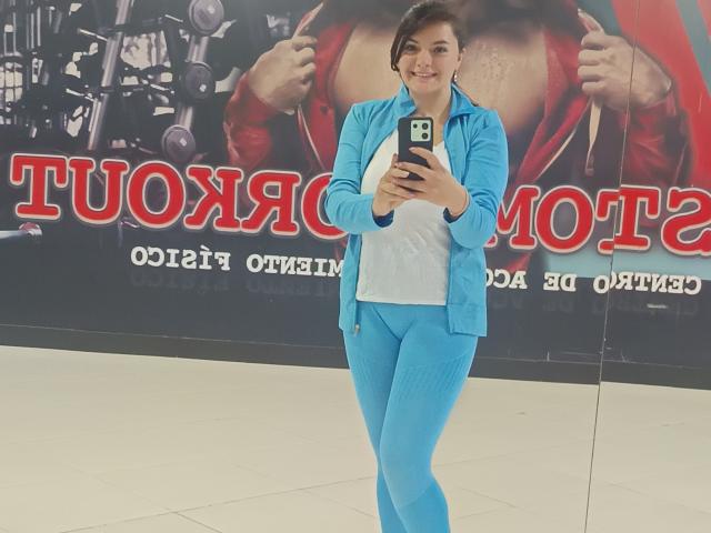 katherinLatin with medium tits, wearing a blue zip-up jacket and matching blue leggings, standing, smiling, holding a phone, taking a selfie, in a bright indoor space.