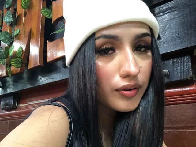 NickiClark has long black hair, a white beanie, and a nose ring. She's looking at the camera with full, glossy lips and dark, thick eyebrows. She's wearing a black top.