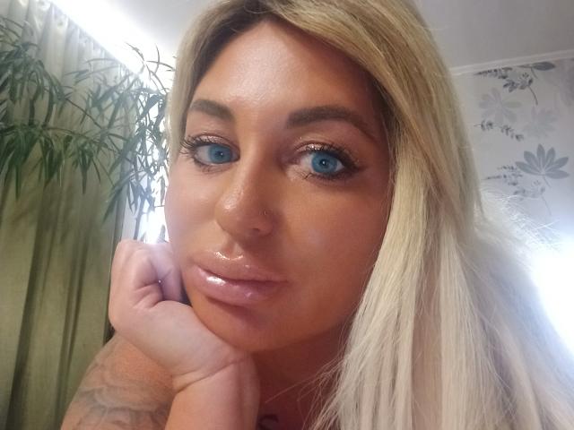 MiraMelody with shiny blue eyes, blonde hair, and glossy pink lips, resting her chin on her hand. Her skin is tan, and she has a nose ring.