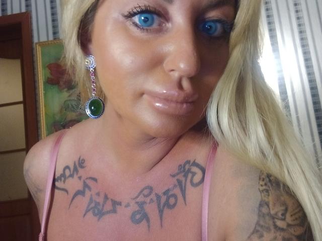 MiraMelody has blue eyes, shiny lips, blonde hair, green earrings, tattoos on her chest, and is wearing a pink top, showing her tits and tattoos.
