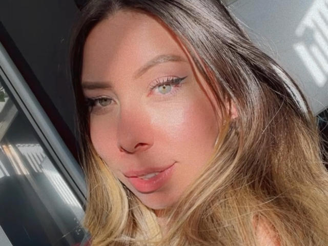 JennyLawry has light brown hair with blonde tips, green eyes, and subtle eyeliner. She's smiling with sunlit skin and natural makeup. Her boobs are not visible.