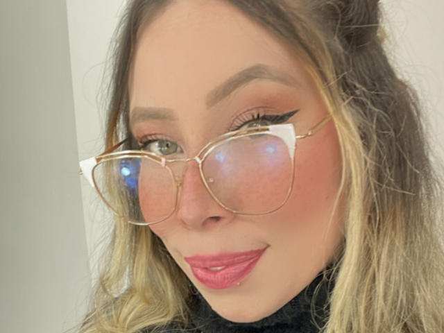 JennyLawry with glossy pink lips, glasses reflecting light, and blonde hair, wearing a black turtleneck. She has winged eyeliner and a slight smile.
