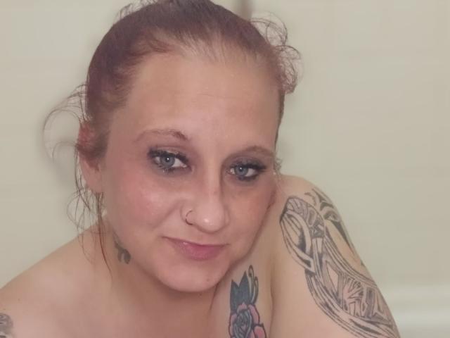 Moonlightluv has light skin, red hair in a messy bun, blue eyes, a nose ring, and tattoos on her shoulder and upper arm. She's topless with a subtle smile.