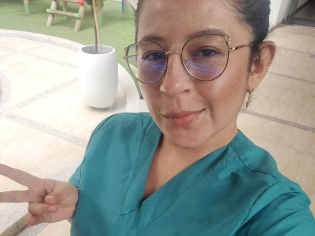 SweetyHot69 with glasses, green shirt, and earrings, smiling, showing a peace sign, and wearing subtle makeup, with a slight blush and visible skin texture.