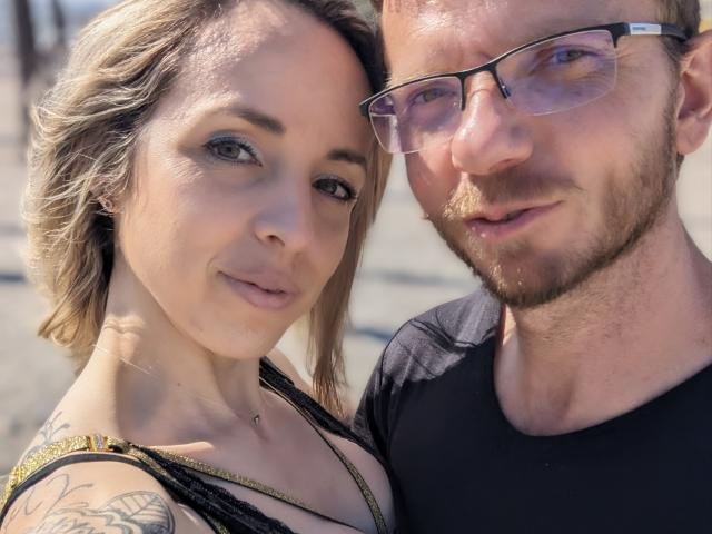 Annic, avec ses cheveux blonds, ses tatouages sur l'épaule, son petit piercing au nez et sa poitrine visible, est assise à côté d'un homme barbu, portant des lunettes et un t-shirt noir..