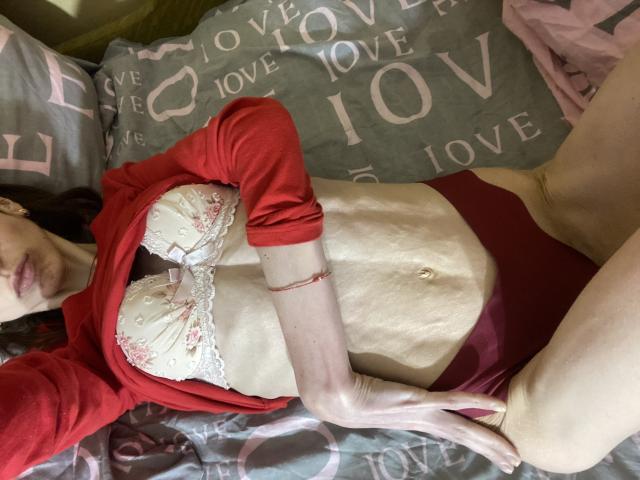 Kabblasha lying on bed, wearing red long-sleeve shirt, white lace bra with pink flowers, red panties, exposing flat, pale belly, right hand on thigh, left arm raised.