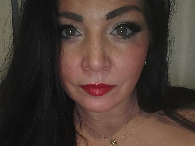 Paiday's face, brown eyes, thick black eyebrows, red lipstick, long black hair, slight freckles, necklace with small pendant, fair skin, looking straight at camera, slightly parted lips.