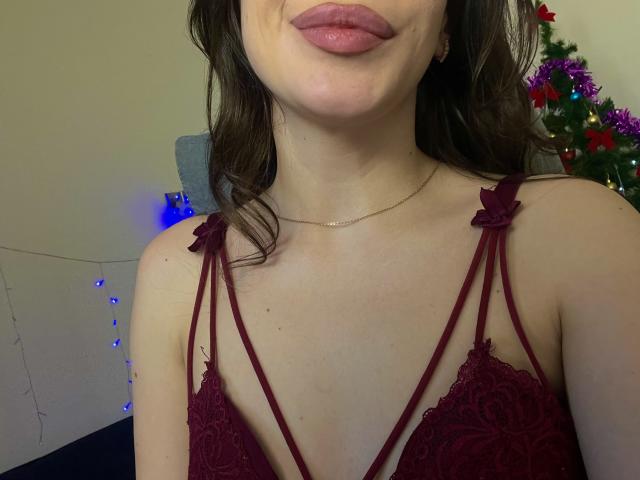 El rostro de Sevillia, mostrando labios rosados, lleva un sujetador de encaje rojo oscuro con detalles de tiras, cabello ondulado castaño, un collar dorado y un tono de piel claro. Se ven los senos a través del encaje..