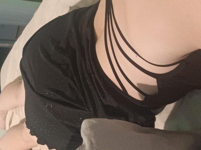MissClaire69 wearing a black, strappy, sparkly dress, exposing her tits through multiple thin straps, lying on a bed with her ass slightly visible.