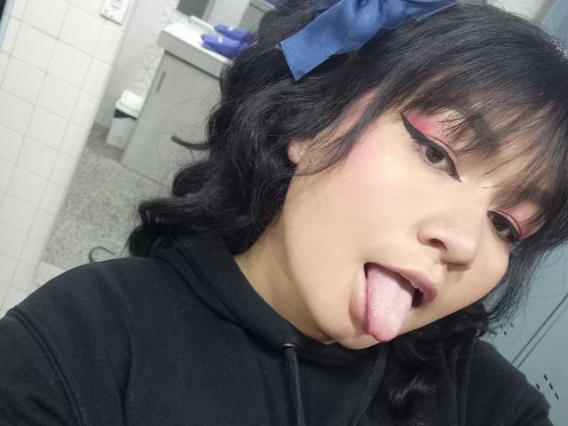 LillyMayer, com cabelo preto ondulado, um laço azul, delineador rosa e moletom preto, língua de fora, olhando para a câmera, em um banheiro, usando maquiagem, mostrando sua língua rosa..