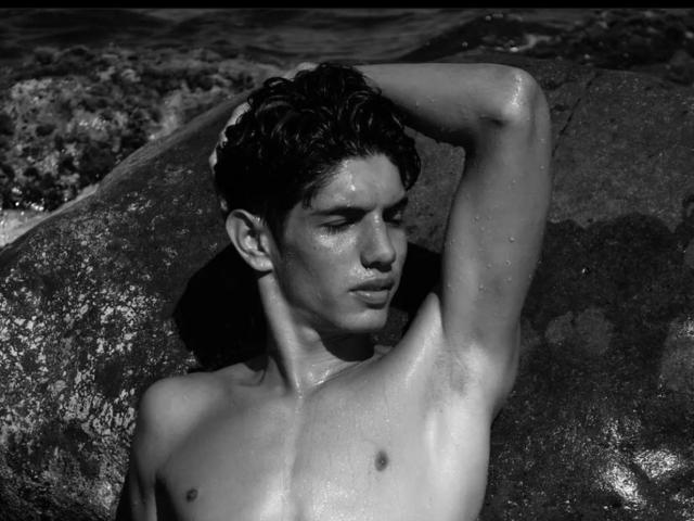 FabrizzioHot with wet, glistening skin, raising arm, showing toned ass, and dark, wavy hair, leaning against rock, in black-and-white shot, looking down, with water droplets.