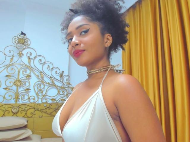 MeganWlliams with curly black hair, red lipstick, and white halter top, showing her tits, wearing a gold chain necklace, standing in front of yellow curtains.