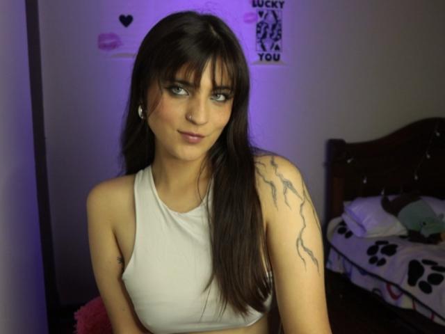 SarahhTanyas, con cabello castaño largo, ojos verdes, top corto blanco que muestra sus pechos, tatuaje en el brazo derecho, sentada en una habitación con poca luz, aro pequeño en la nariz, ligera sonrisa..