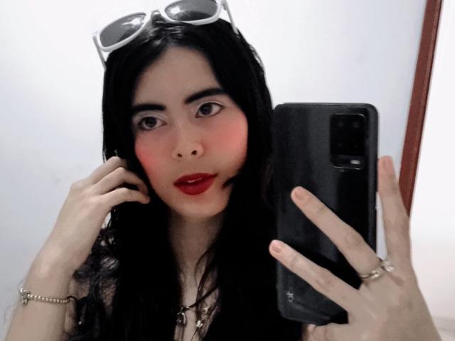TiffanyBaez with black hair, red lipstick, and eyeglasses on her head, taking a selfie with her phone, showing off her blushed cheeks and ring on her right hand.