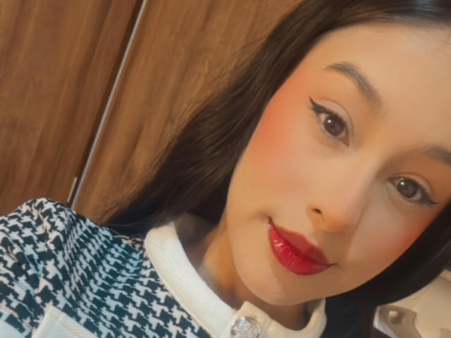 Soranaa has red lips, black eyeliner, and a slight smile. She's wearing a houndstooth shirt with a white collar, and has long black hair.