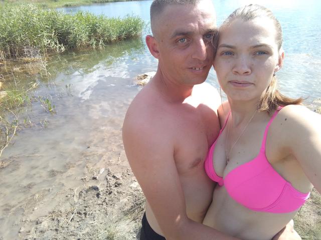 Shirtless ParaFox with short brown hair, tan skin, and small tits stands behind blonde ParaFox in pink bra, by water. She has blue eyes, he has a slight smile.