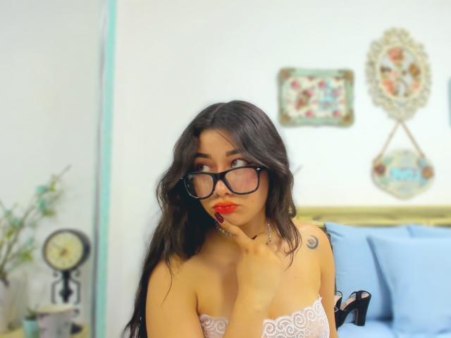 Genesiss with dark hair, glasses, red lipstick, and white lace top, touching her chin, showing a small crescent moon tattoo on her left shoulder, looking up.