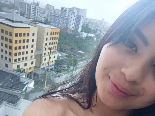 EmmaaRodriguez with puffy lips, dark brown hair, and a slight smile, showing her bare shoulders, in a cityscape with yellow and white buildings.