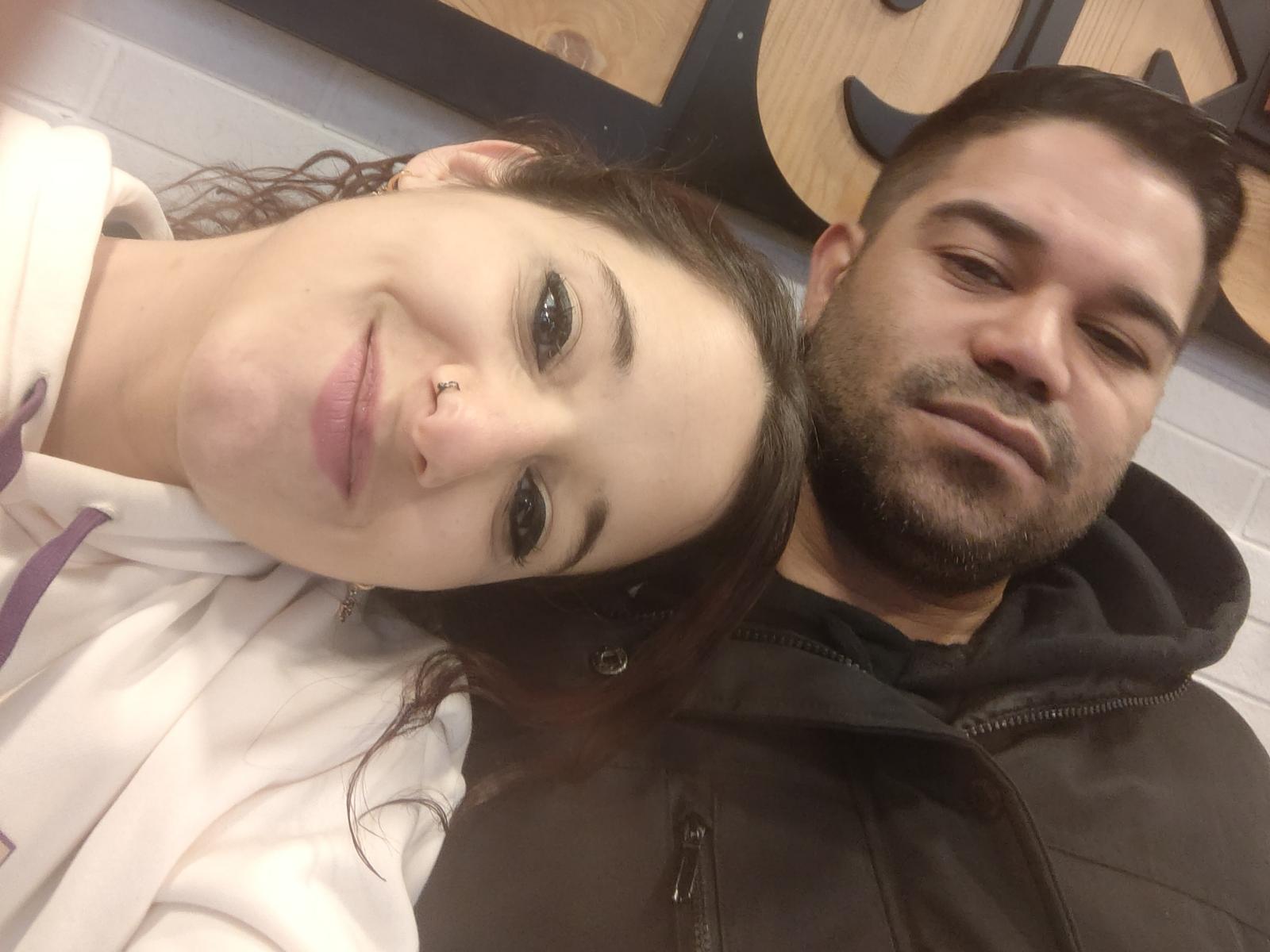 Gustosos with nose ring, smiling, and brown curly hair, next to Gustosos with black beard and short hair, in black hoodie, both looking at camera, in selfie.