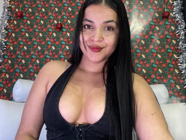 HikariChloe has long black hair, red lipstick, and a black halter top showing her big tits. She's smiling with a slight nose piercing and silver tinsel in the background.