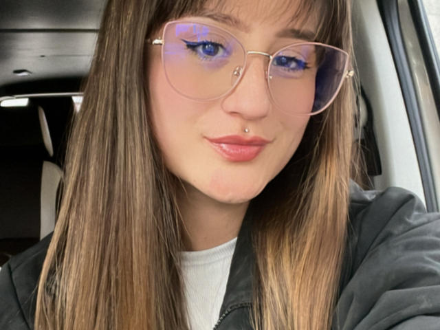 NinaBryons with blue eyes, wearing pink glasses, has a nose piercing and a lip piercing. She's got long brown hair, a black jacket, and a white top.