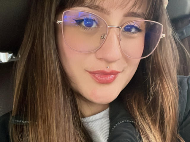 NinaBryons has blue eyes with reflective glasses, a nose piercing, and glossy red lips. She's wearing a black jacket over a white turtleneck, with brown hair.