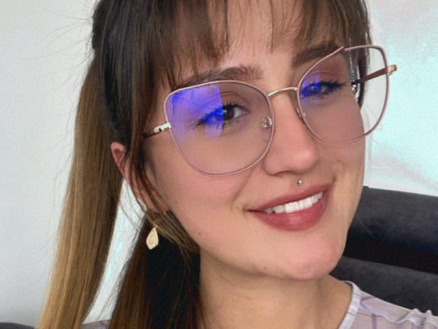 NinaBryons with puffy blue-tinted glasses, brown ponytail, nose piercing, and lip piercing, smiling at the camera. She's got a cute face and small tits.
