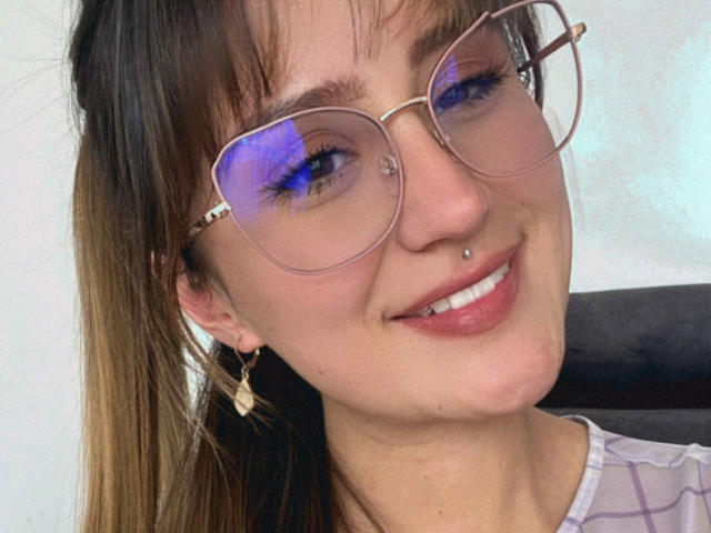 NinaBryons has blue eyeshadow, a nose ring, and a lip piercing. She's smiling with her glasses on, wearing a striped shirt and gold earrings.