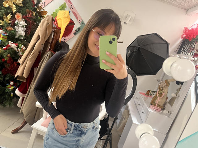 NinaBryons with long brown hair, glasses, black turtleneck, blue jeans, green phone, smiling, holding phone in mirror selfie, black umbrella in background, Christmas tree on left.