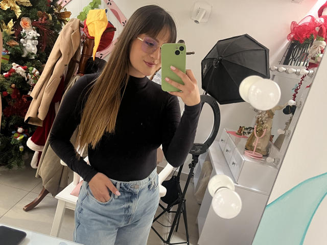 NinaBryons with long brown hair, glasses, black turtleneck, and light blue jeans, taking a mirror selfie with her green phone, showing her cute ass in the reflection.