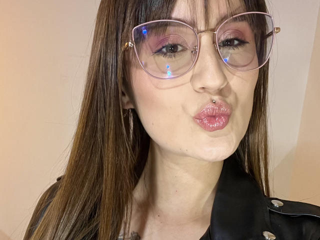 NinaBryons with puffy pink lips, brown hair, and glasses, making a kissing face. She has a nose piercing and wears a black leather jacket.