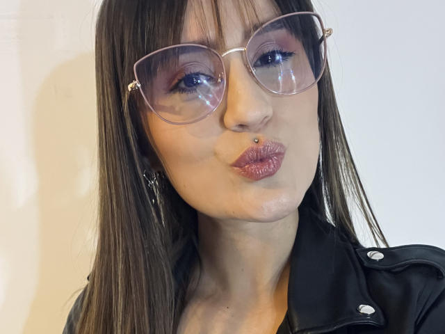 NinaBryons with puffy pinkish-purple lipstick, brown straight hair, and clear glasses, pouting her glossy lips, wearing a black leather jacket, and a nose piercing.