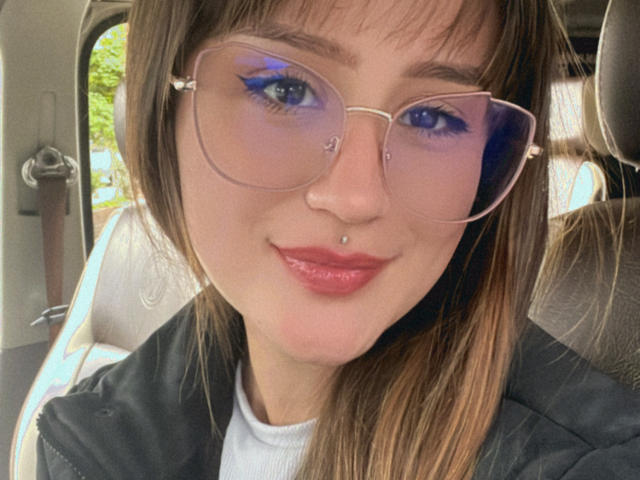 NinaBryons has blue eyeshadow, a nose piercing, and a lip piercing, wearing glasses with a heart-shaped top. Her ass is not visible. She's in a car.