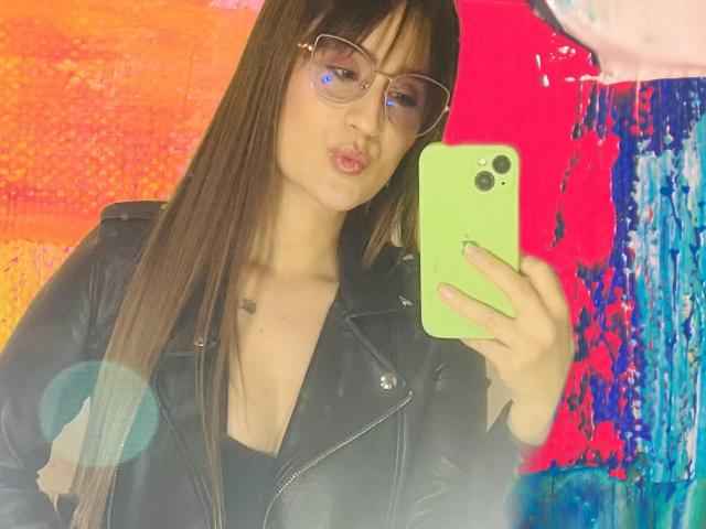 NinaBryons with glasses, pouty lips, and long brown hair, wearing a black leather jacket, holding a green phone, reflecting in a mirror. Her ass is not visible.