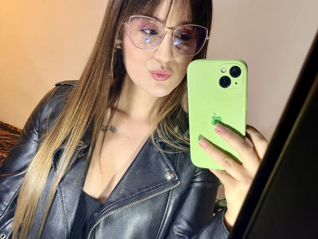 NinaBryons with long brown hair, wearing glasses, a black leather jacket, and a green iPhone, taking a mirror selfie, showing her pouty lips and small tits.