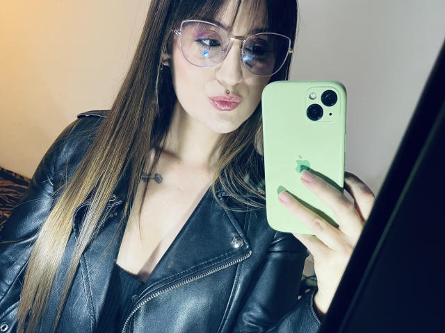 NinaBryons with glasses, holding a green iPhone, wearing a black leather jacket, showing her cleavage, taking a mirror selfie, her lips puckered, her long brown hair, small tattoo.