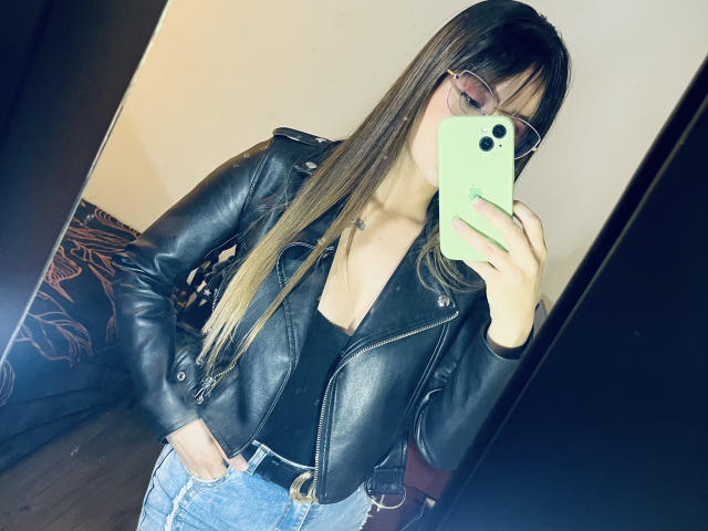 NinaBryons with long brown hair, glasses, black leather jacket, low-cut black top, blue jeans, holding green phone, mirror selfie, showing her tits, in a room.