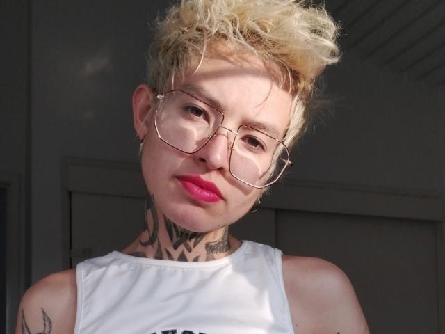 Lilyanna69 with short blonde curly hair, glasses, pink lipstick, white tank top, tattooed neck, and a confident, slightly smirking expression.