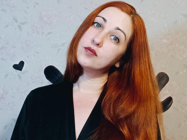 JillSilvies has red hair, blue eyes, and is wearing a black velvet robe. Her pouty lips are slightly glossy. Black wing decorations are on her robe.
