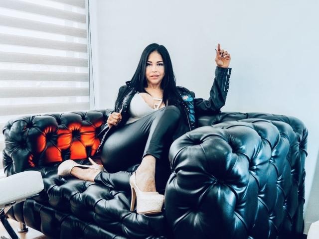 AngelinaDeleon with black hair, sitting on a tufted black leather couch, wearing a black jacket, grey top, black pants, and beige heels, holding a cigarette, giving a peace sign.