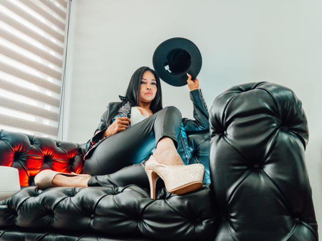 AngelinaDeleon lounging on a black tufted leather couch, holding a black hat, wearing grey pants, a blue shirt, and pink high heels, with a confident, sexy look.