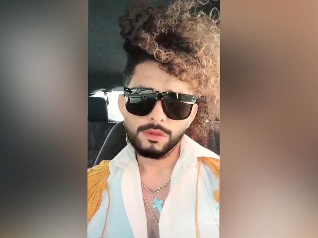 Dandann with curly blonde and brown hair, black sunglasses, beard, white jacket with yellow epaulets, necklace, and a blue star pendant, looking serious in a car.