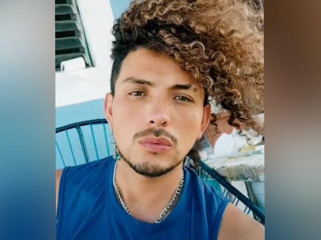 Dandann with curly brown hair, medium skin, and a goatee, wearing a blue sleeveless shirt and a silver chain, looking straight at the camera with a neutral expression.