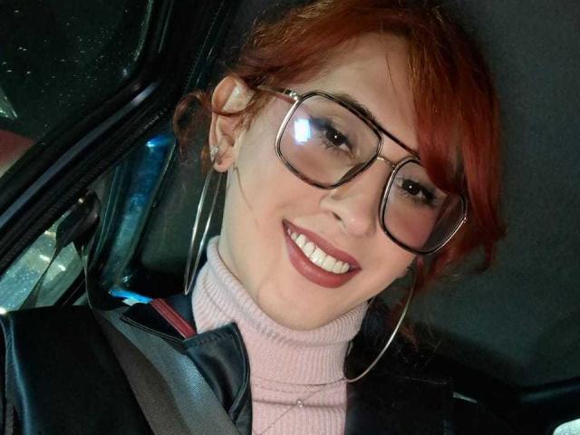 JulianaSaenz with red hair, glasses, and a smile, wearing a pink turtleneck, black leather jacket, and seatbelt, looking cute with visible nose ring and ear piercings.