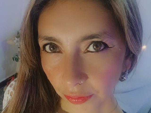 MichelleDumont's face with brown eyes, long brown hair, red lipstick, nose piercing, and winged eyeliner, wearing small earrings, looking at the camera with a slight smile.