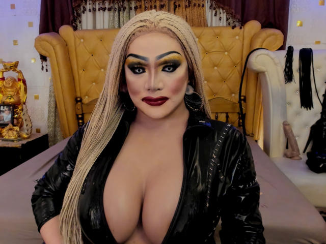 QueenOfPainSasha with huge tits, blonde braids, black leather jacket, deep cleavage, bold yellow eyeshadow, dark red lipstick, standing in front of a yellow tufted headboard.