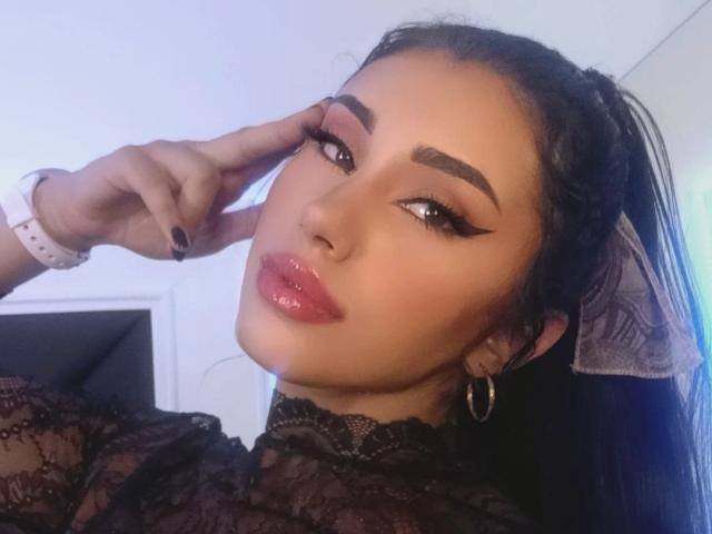 YennettSphinx with glossy pink lips, dark eyebrows, long black eyeliner, and a pink wristwatch, touching her cheek with black nails, wearing lace and a grey hair ribbon.